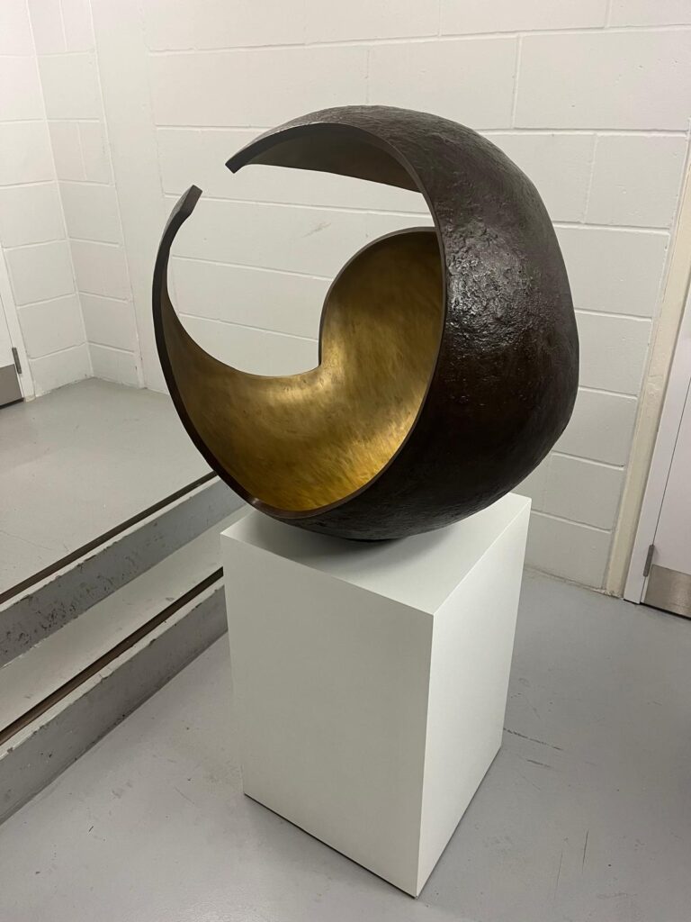 Abstract sculpture titled 'Enclose' by Vera Borghgraef, featuring a circular form with a textured dark exterior and a smooth gold interior, displayed on a white pedestal.