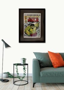 Framed Incredible Hulk comic book cover displayed on a wall above a sofa with orange cushion and a side table with a plant.