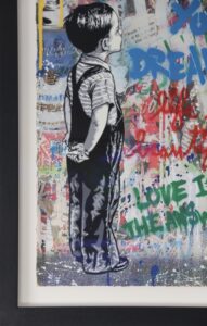 Boy in striped shirt and overalls with hands behind back standing against a colourful graffiti wall