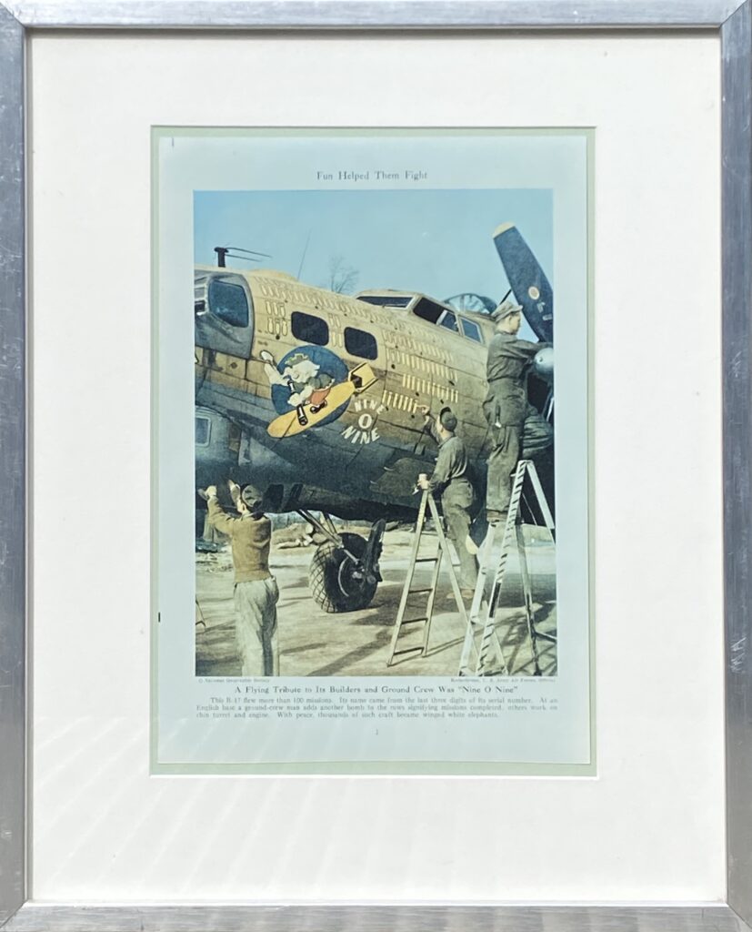 Framed artwork featuring an airplane with cartoon-style artwork on its nose, surrounded by people performing maintenance.