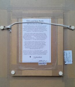 Back view of a framed artwork titled 'Surging Waters III' by artists Chris and Steve Rocks, featuring hanging wire and stickers.